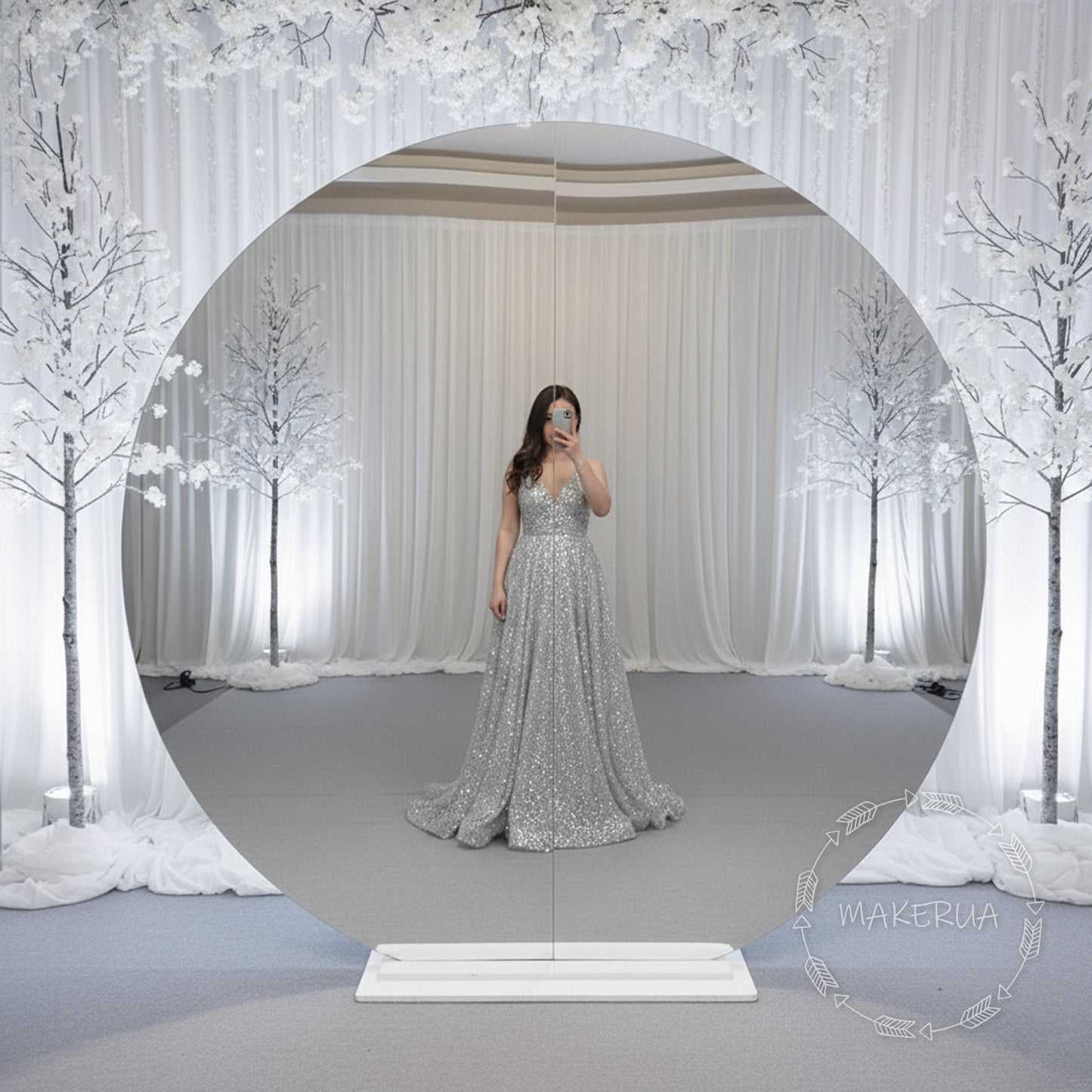 Woman in a silver dress standing in front of a circular mirror backdrop with a decorative background.