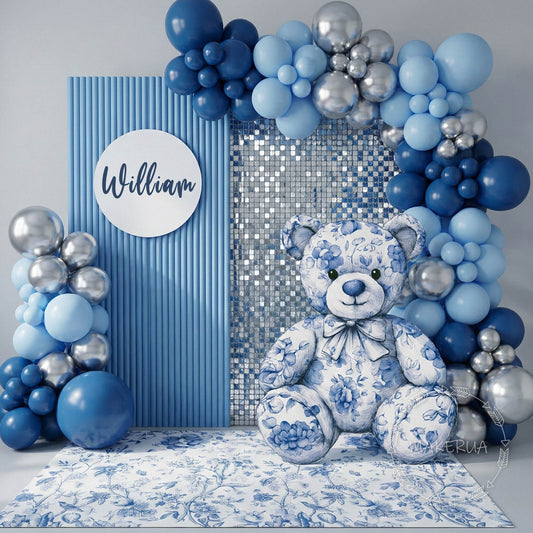 Decorative setup with blue and silver balloons, a cutout dior teddy bear, and a name plaque on a floral-patterned floor.