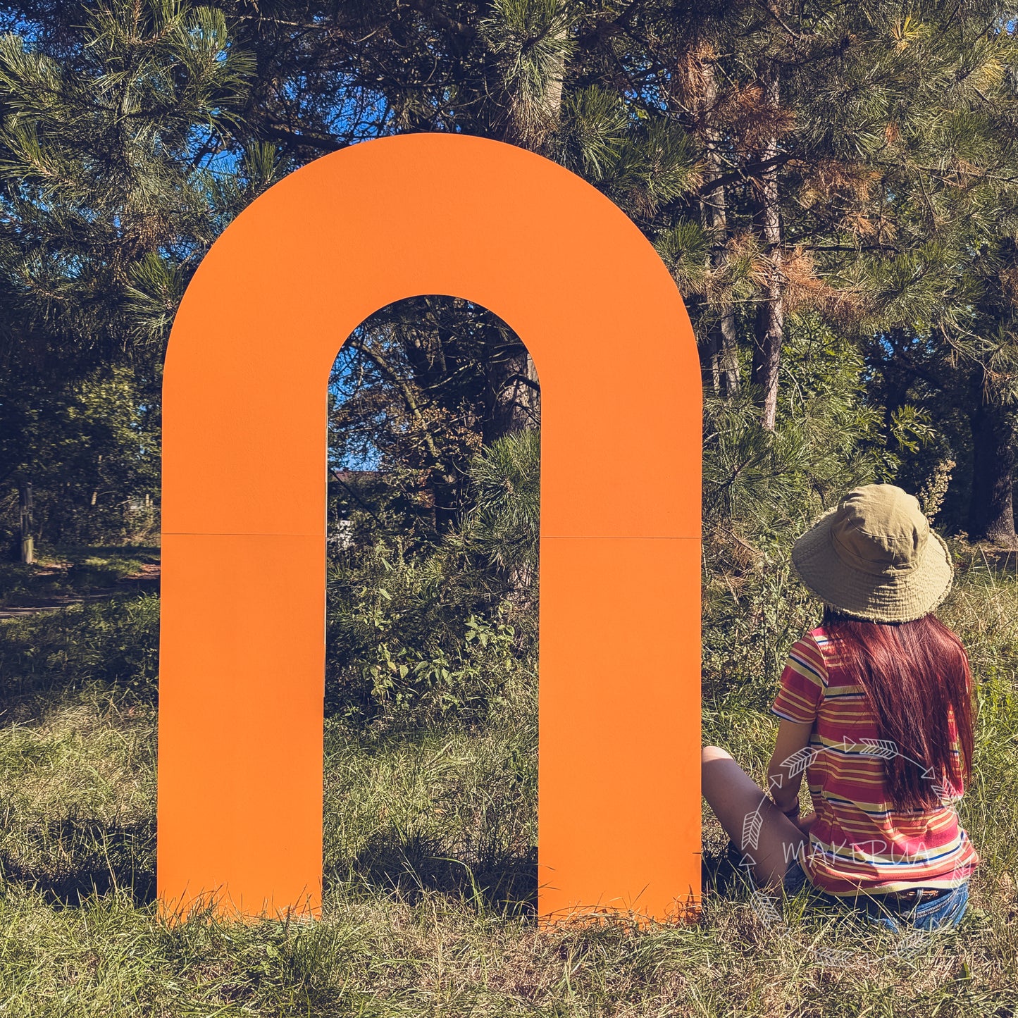 Large, thick U-shaped solid arch painted vibrant orange, standing outdoors in a grassy park setting next to a person for scale. Trendy modern block backdrop for colorful boho weddings, festival photo booths, and outdoor events.