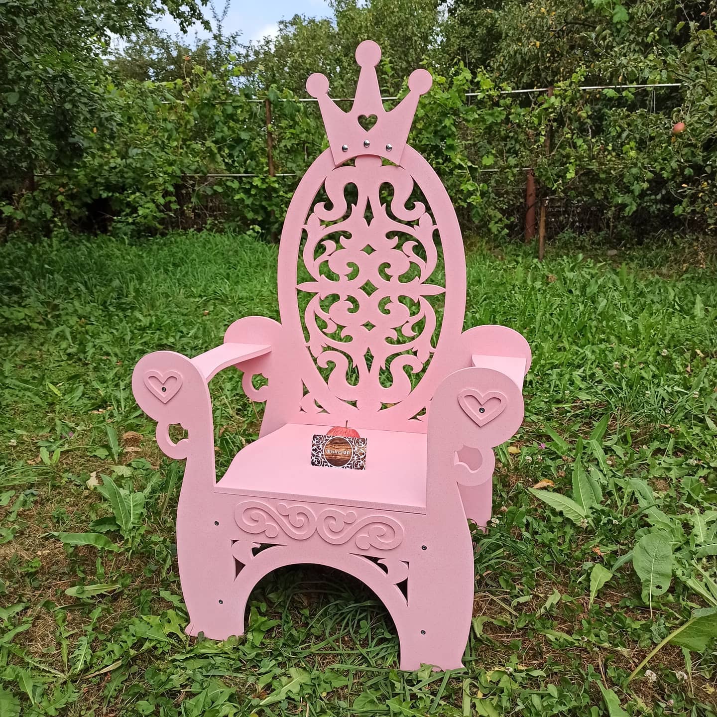 Pink wooden princess chair featuring a detailed filigree backrest and heart-shaped accents on the armrests. Photographed outdoors on green grass. Beautiful fairytale photo prop for girls' birthday parties and outdoor summer events.
