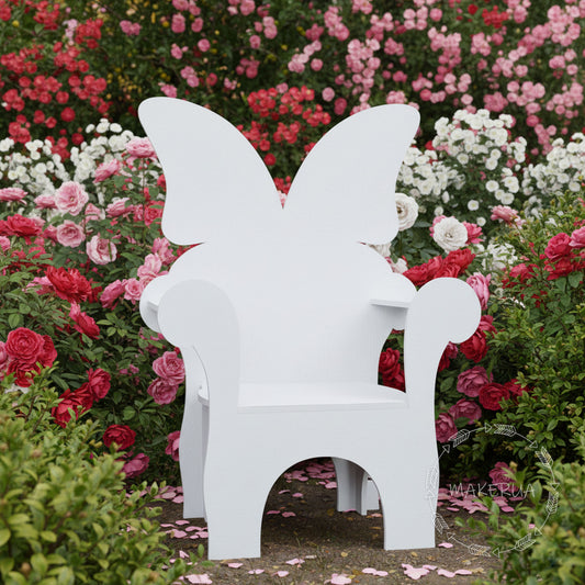 White wooden kids chair featuring a large butterfly wings backrest design. Placed outdoors in a beautiful garden with pink blooming roses. Enchanted fairytale photo prop for girls' birthday parties and spring photo shoots.