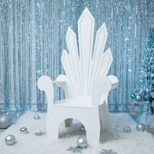 White throne chair in a snowy scene with silver ornaments and a sparkling blue curtain.